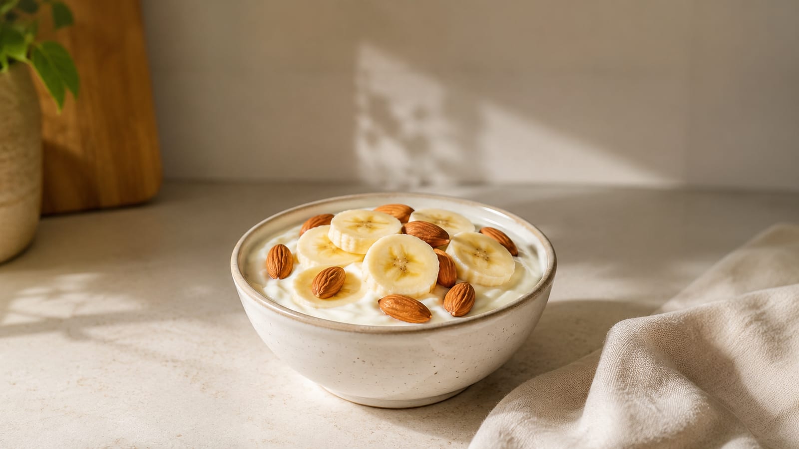 Ceramic bowl of yogurt topped with banana slices and almonds on a sunlit counter